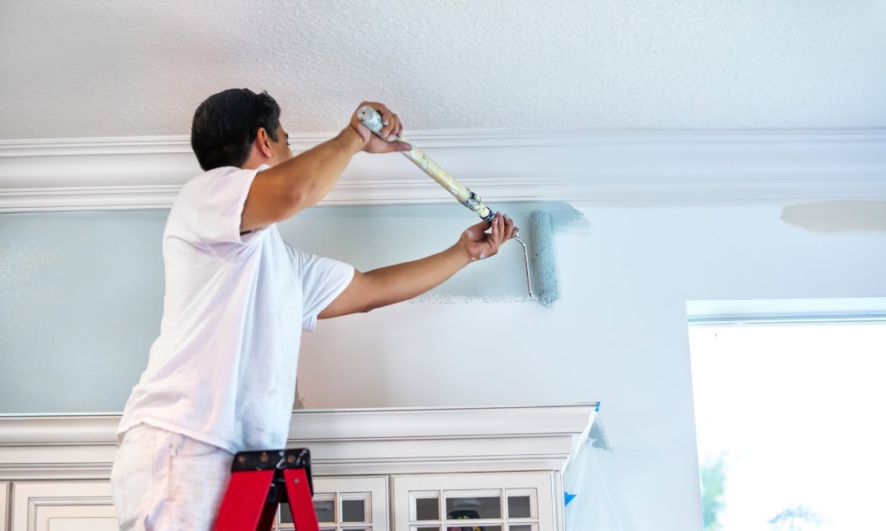 Person painting a light blue wall on a red ladder in a bright room.